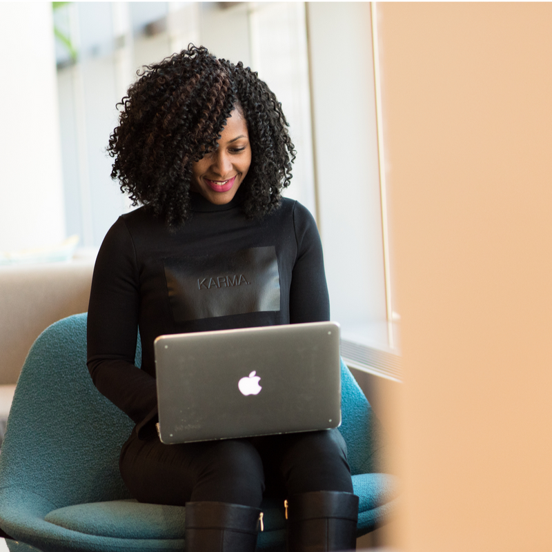 Black woman in a teal chair working on a Mac Book. Woman is smiling, wearing black clothes and in front of a window. 